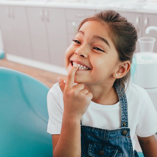 A little girl wearing denim overalls and pointing to her healthy teeth while at the dentist’s office